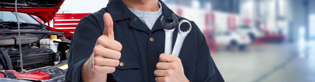 A mechanic in a work uniform gives a thumbs up while holding a wrench and spanner in front of a car engine, signaling a job well done.